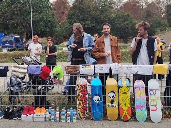 Einige junge Herren stehen hinter einem Zaun, geschmückt mit Skateboards, Getränken und Anderem