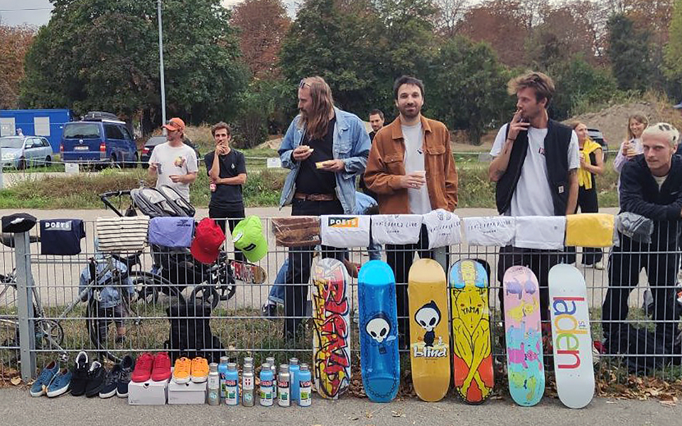 Einige junge Herren stehen hinter einem Zaun, geschmückt mit Skateboards, Getränken und Anderem