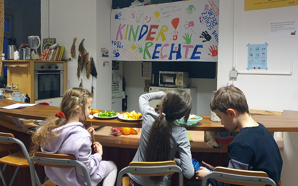 Drei Kinder essen eine Jause mit einem bunten "Kinderrechte" Plakatt im Hintergrund